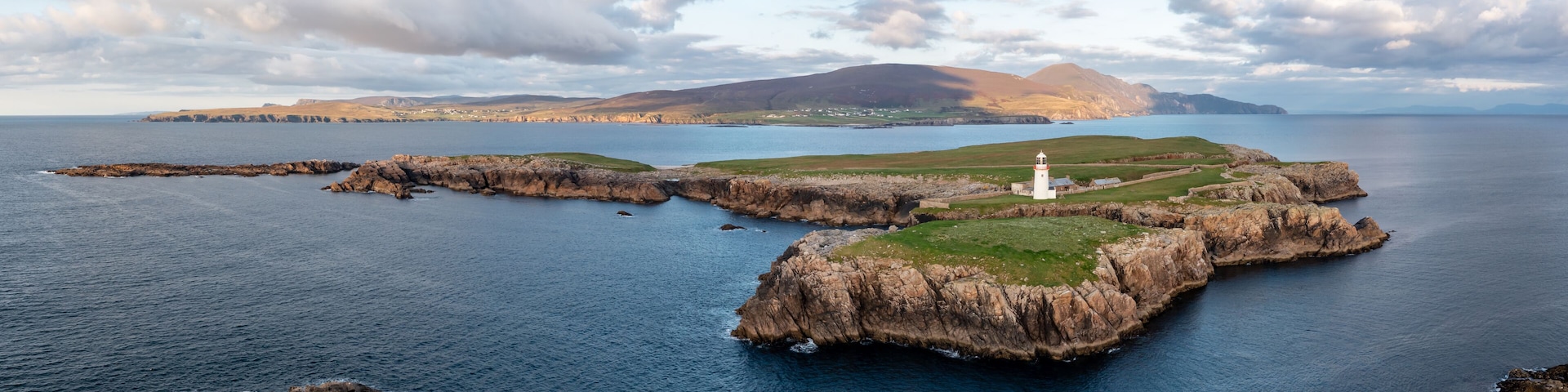 A rugged coastline with a lighthouse, rocky outcrops, and a distant mountain range.