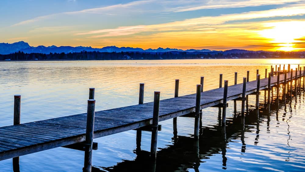 A wooden pier extending into calm waters with a sunset and mountains in the background.