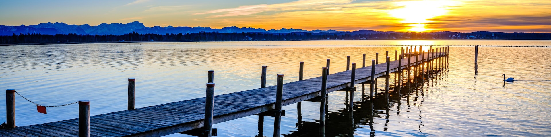 A wooden pier extending into calm waters with a sunset and mountains in the background.