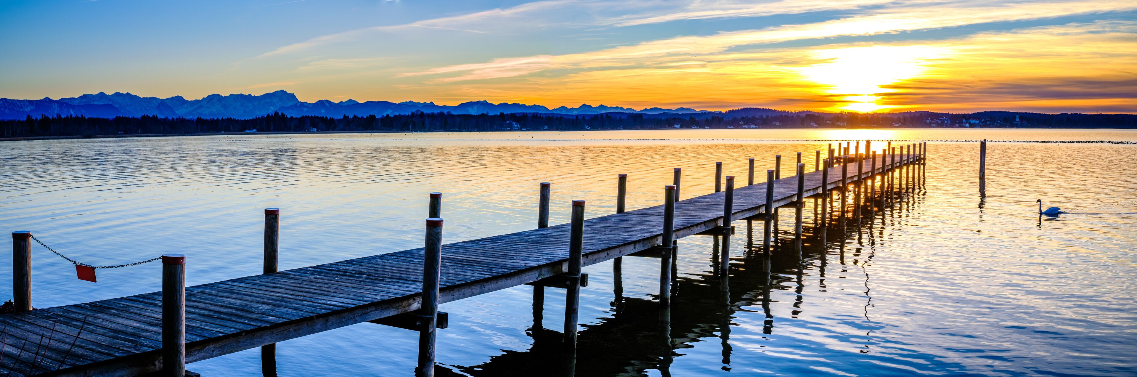 A wooden pier extending into calm waters with a sunset and mountains in the background.