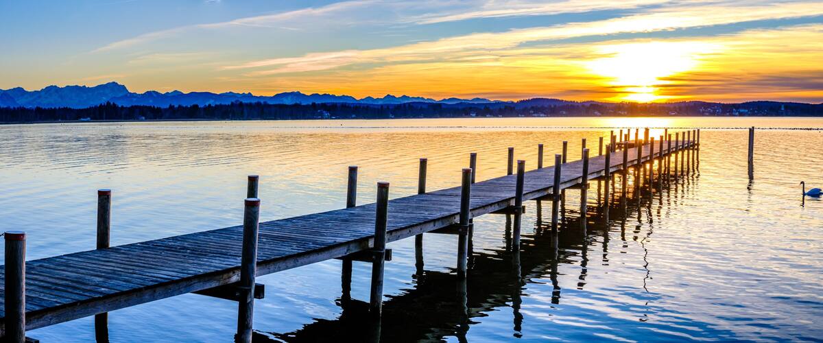 A wooden pier extending into calm waters with a sunset and mountains in the background.