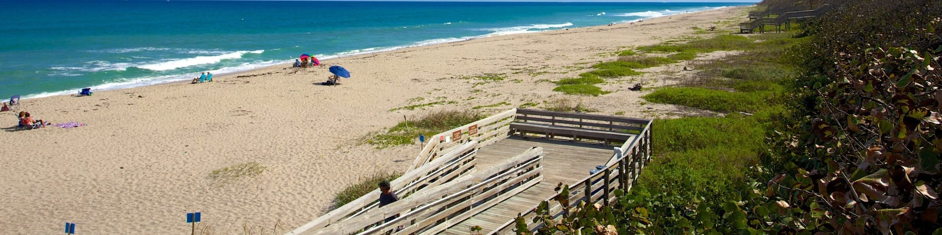 A beach with a wooden boardwalk, people sunbathing, and buildings in the distance.