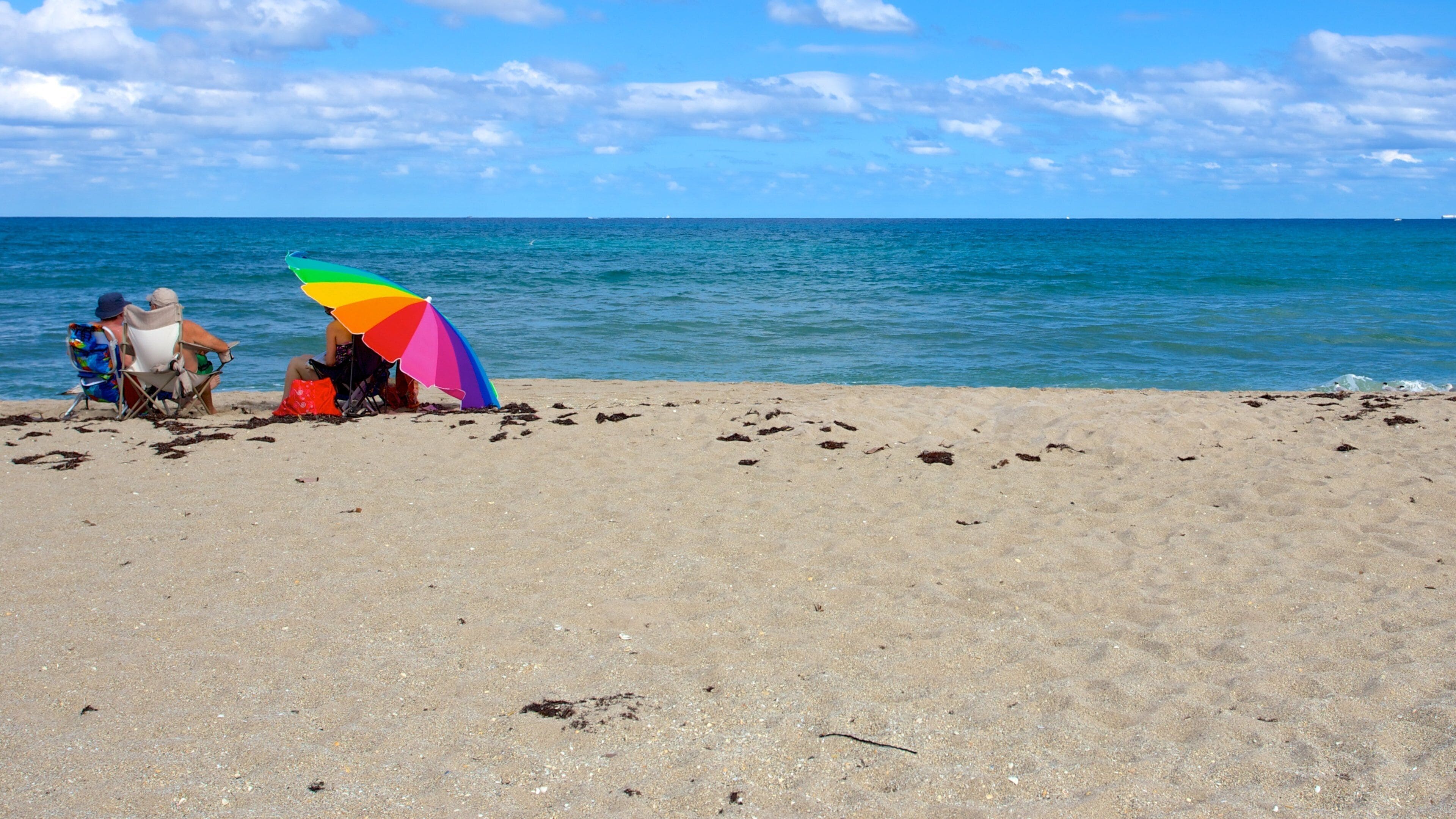 A beach with people sitting under a colorful umbrella.