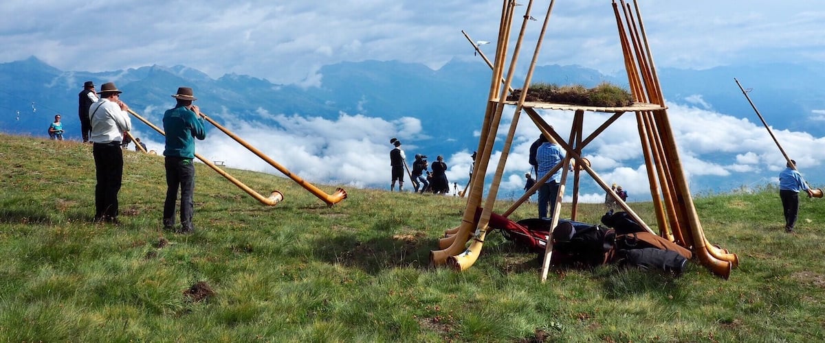 Eine Gruppe von Menschen auf einer grasbewachsenen Anhöhe mit einer Holzkonstruktion und langen Pfählen, mit Blick auf eine Berglandschaft mit darunterliegenden Wolken.