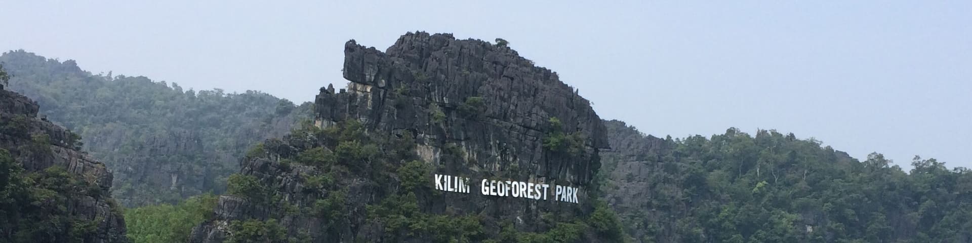 A large rock formation with a sign that reads "Kiling GeoForest Park" surrounded by water and greenery.