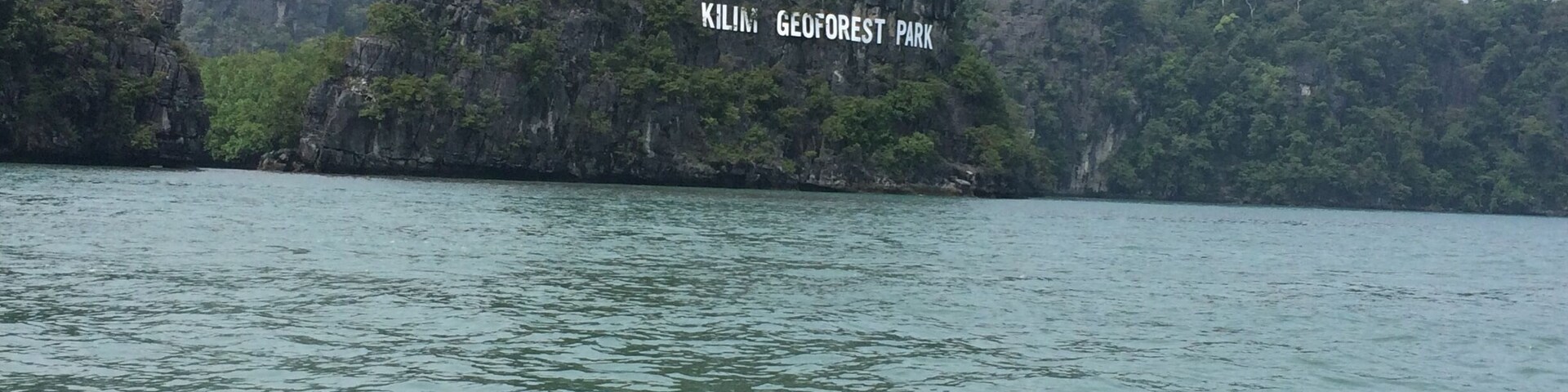 Una gran formación rocosa con un letrero que dice "Kiling GeoForest Park" rodeada de agua y follaje.