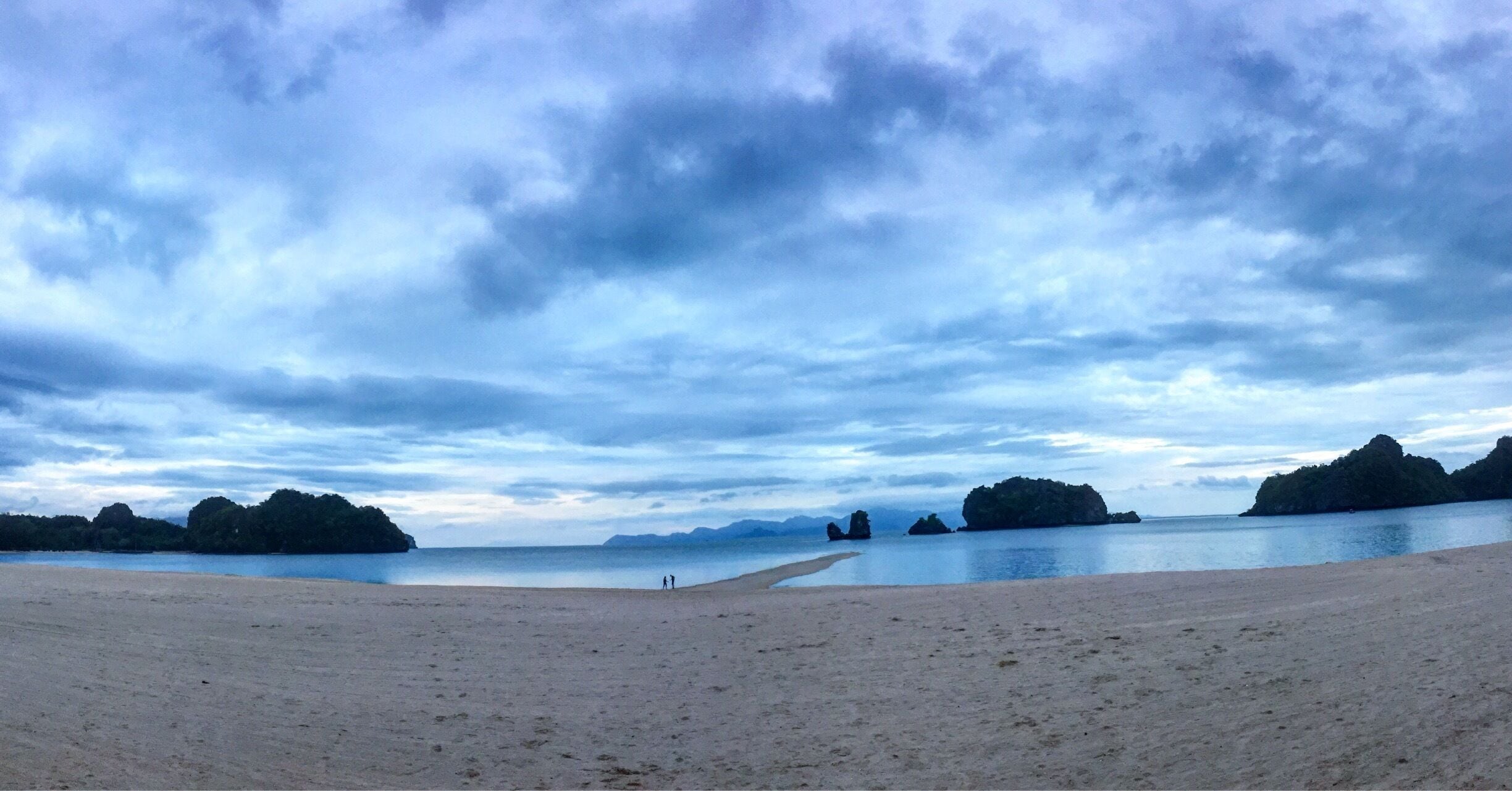 Una playa con orilla arenosa, agua tranquila y unas islas rocosas distantes bajo un cielo nublado.