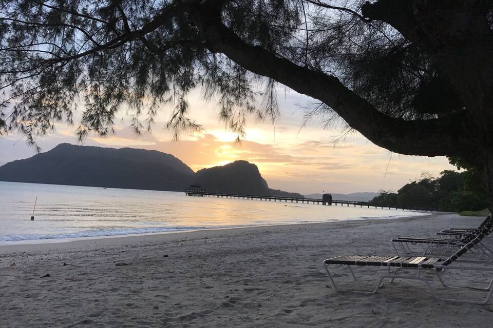 Una platja amb un pont de fusta que s'endinsa en aigües tranquil·les, envoltada de muntanyes i arbres.