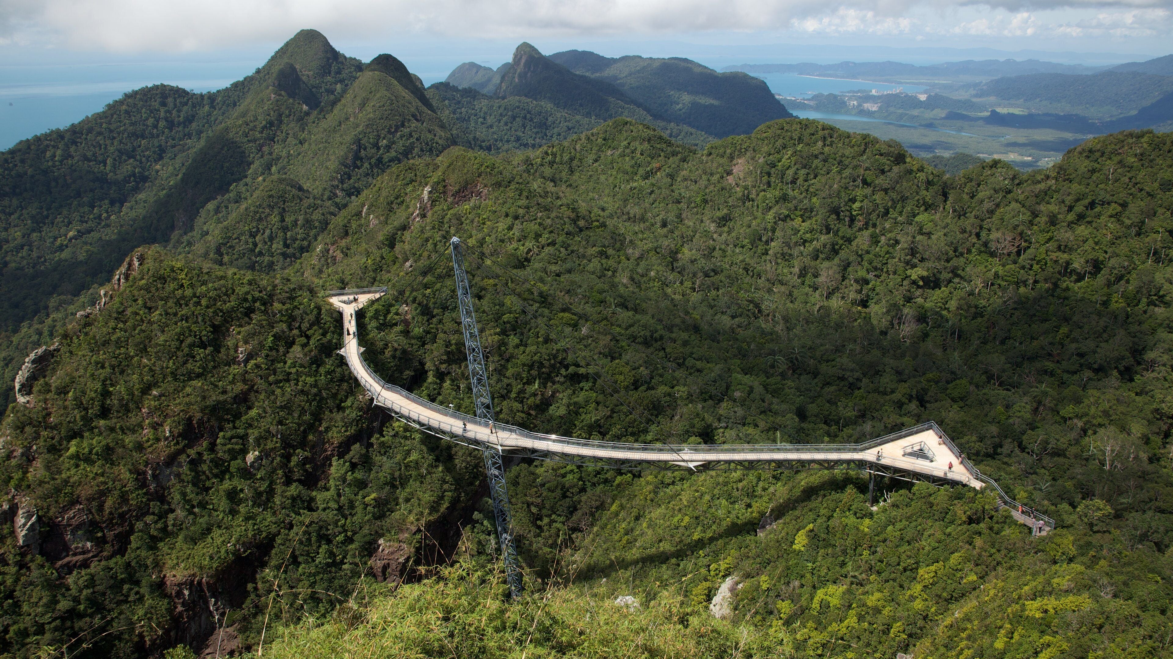 A long bridge curving through a dense forest with mountains in the background.