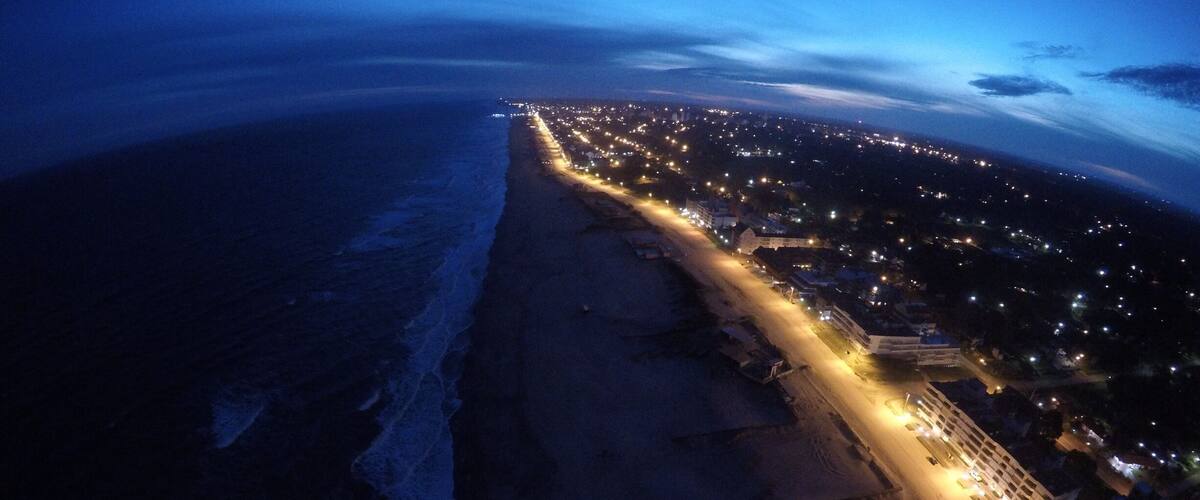An aerial view of a coastal city at dusk with a beach, a lit promenade, and a densely populated area with lights.