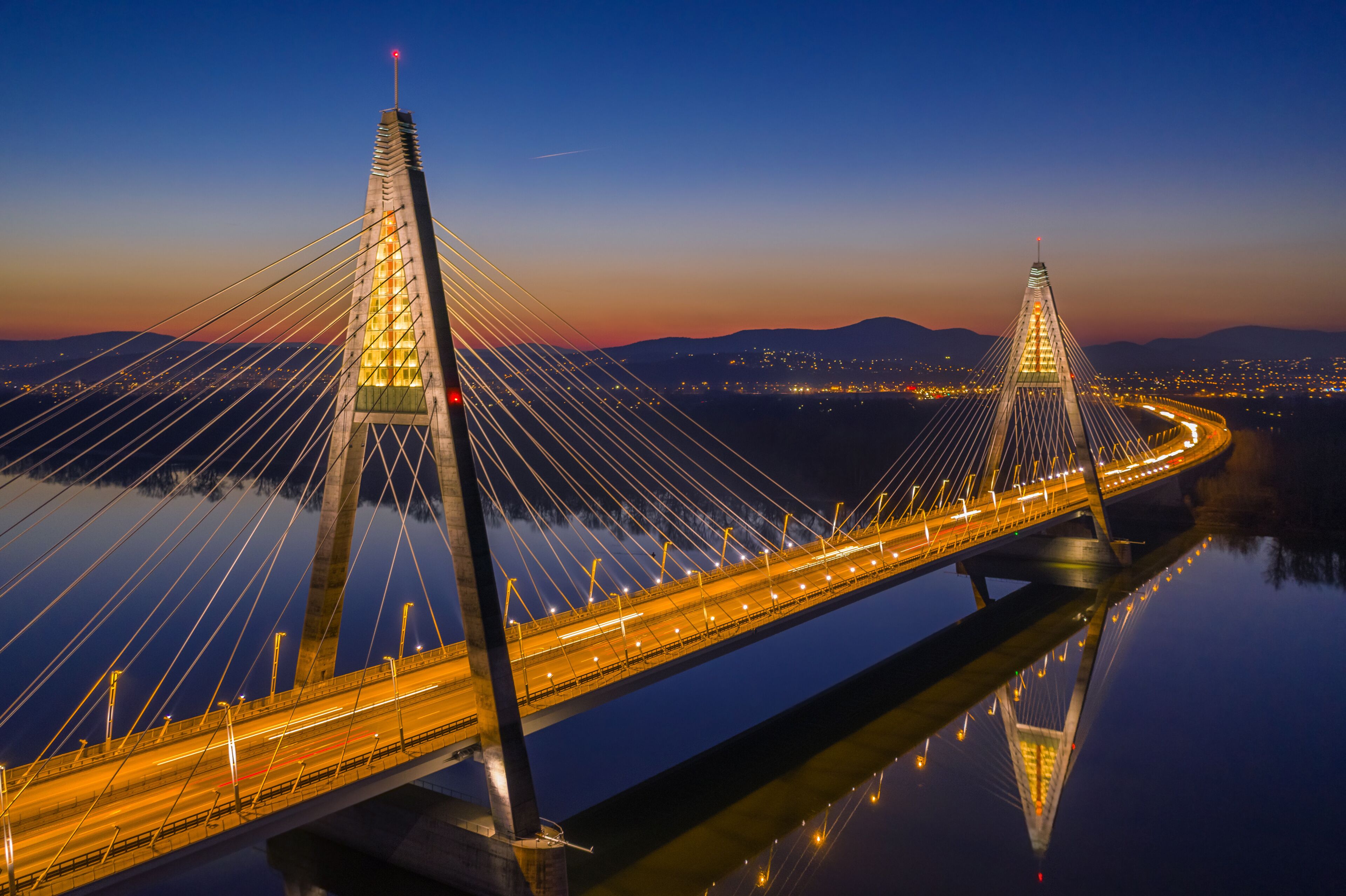 A cable-stayed bridge at twilight with illuminated cables and a clear reflection on the water.