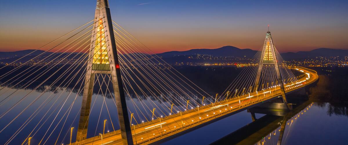 A cable-stayed bridge at twilight with illuminated cables and a clear reflection on the water.