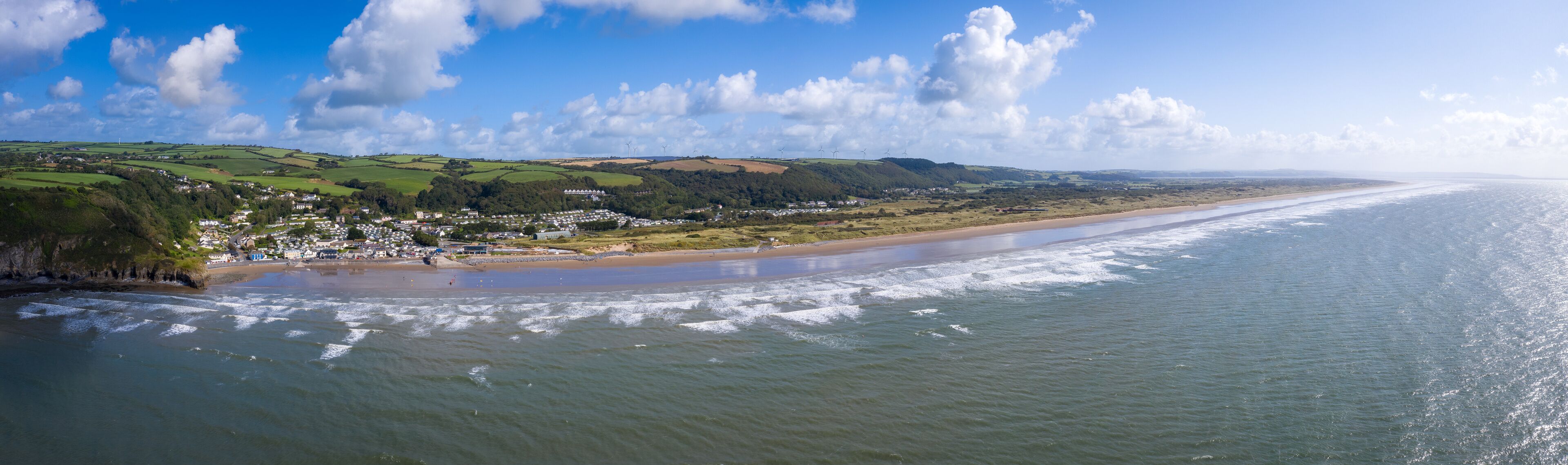 A coastal town with a beach, rolling hills, and wind turbines in the distance.