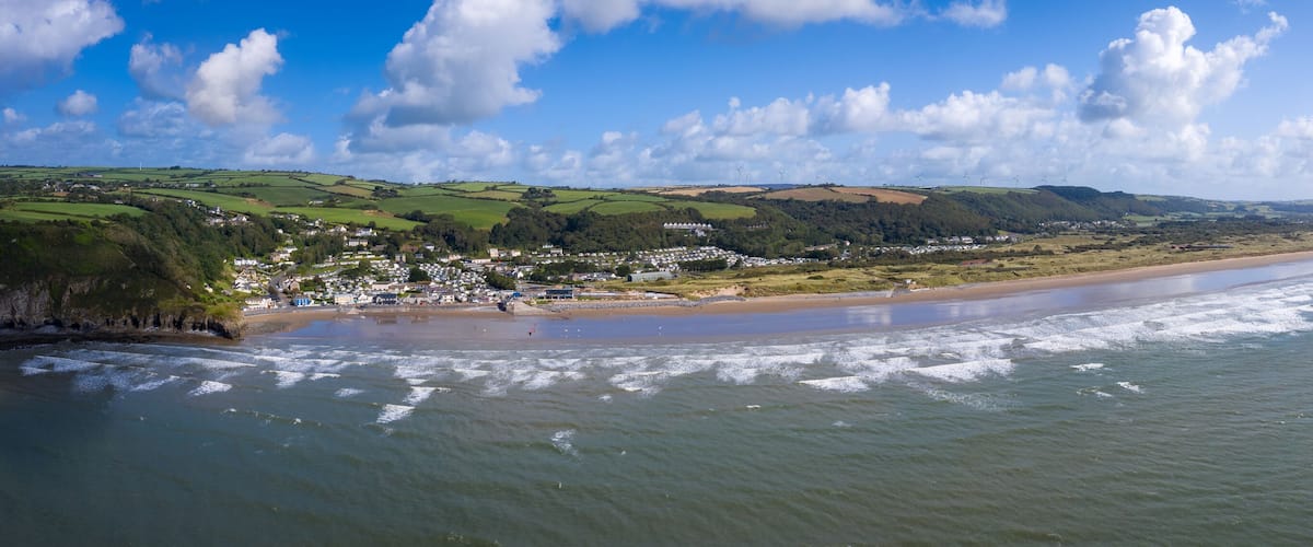 A coastal town with a beach, rolling hills, and wind turbines in the distance.
