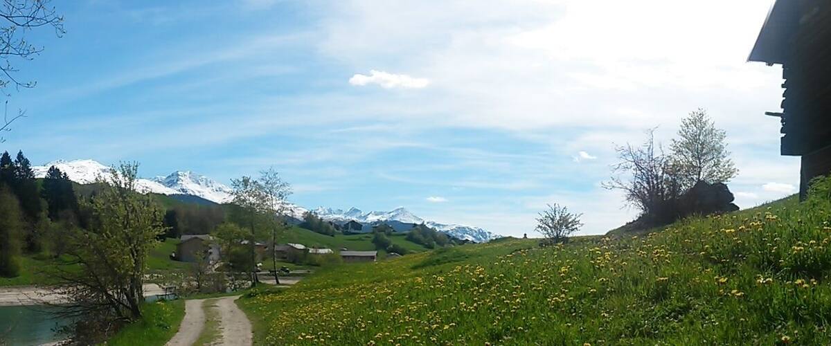 A rural landscape with a river, green fields, a wooden building, and snow-capped mountains in the distance.