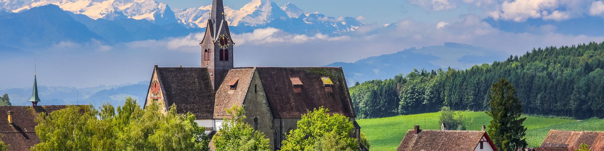 Una iglesia con un campanario alto, rodeada de casas y vegetación exuberante, con montañas nevadas al fondo.