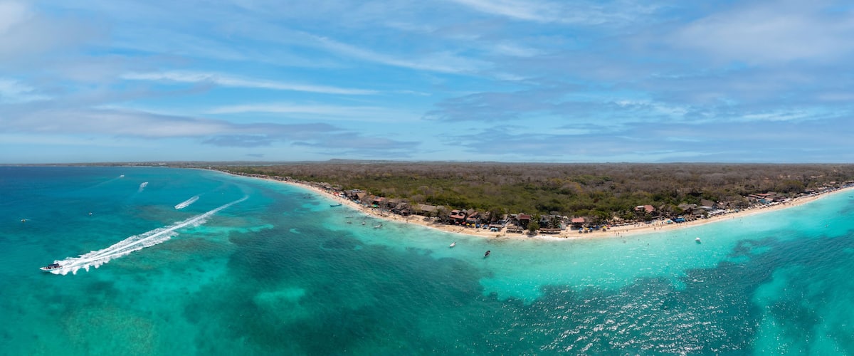 An aerial view of a coastal area with clear turquoise waters, a sandy beach, and a dense forested area.