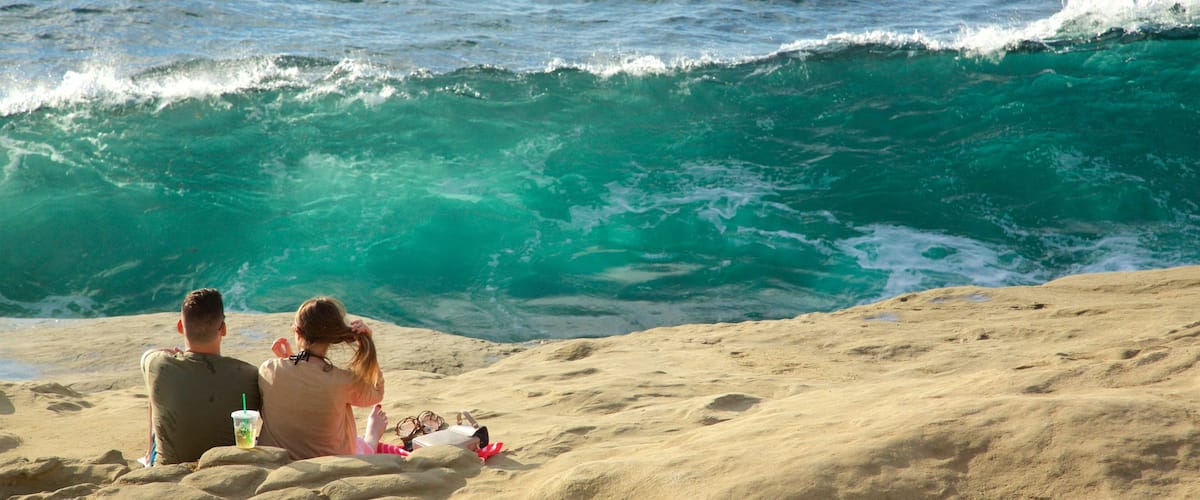 A couple sitting on a sandy beach with a large wave crashing in the background.