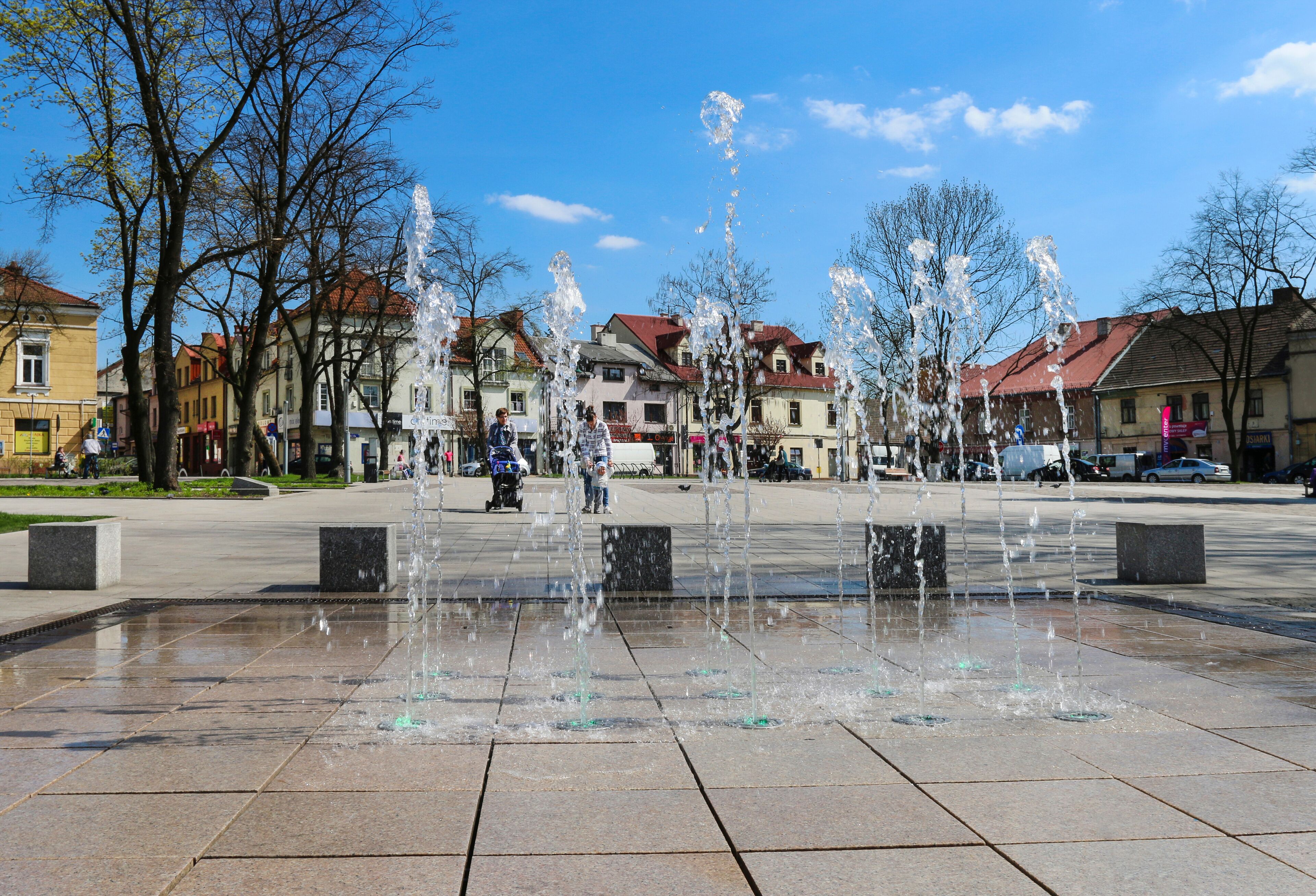 Uma praça pública com uma fonte de água, edifícios e árvores.