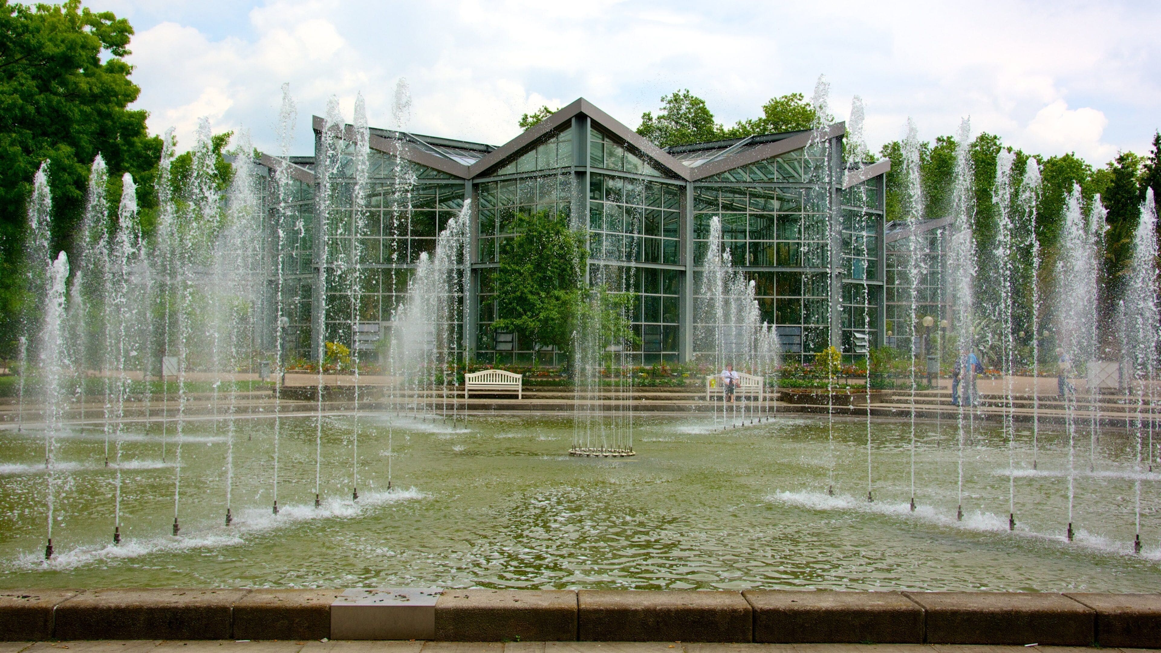 A modern glass building with a triangular roof, surrounded by a water fountain display.
