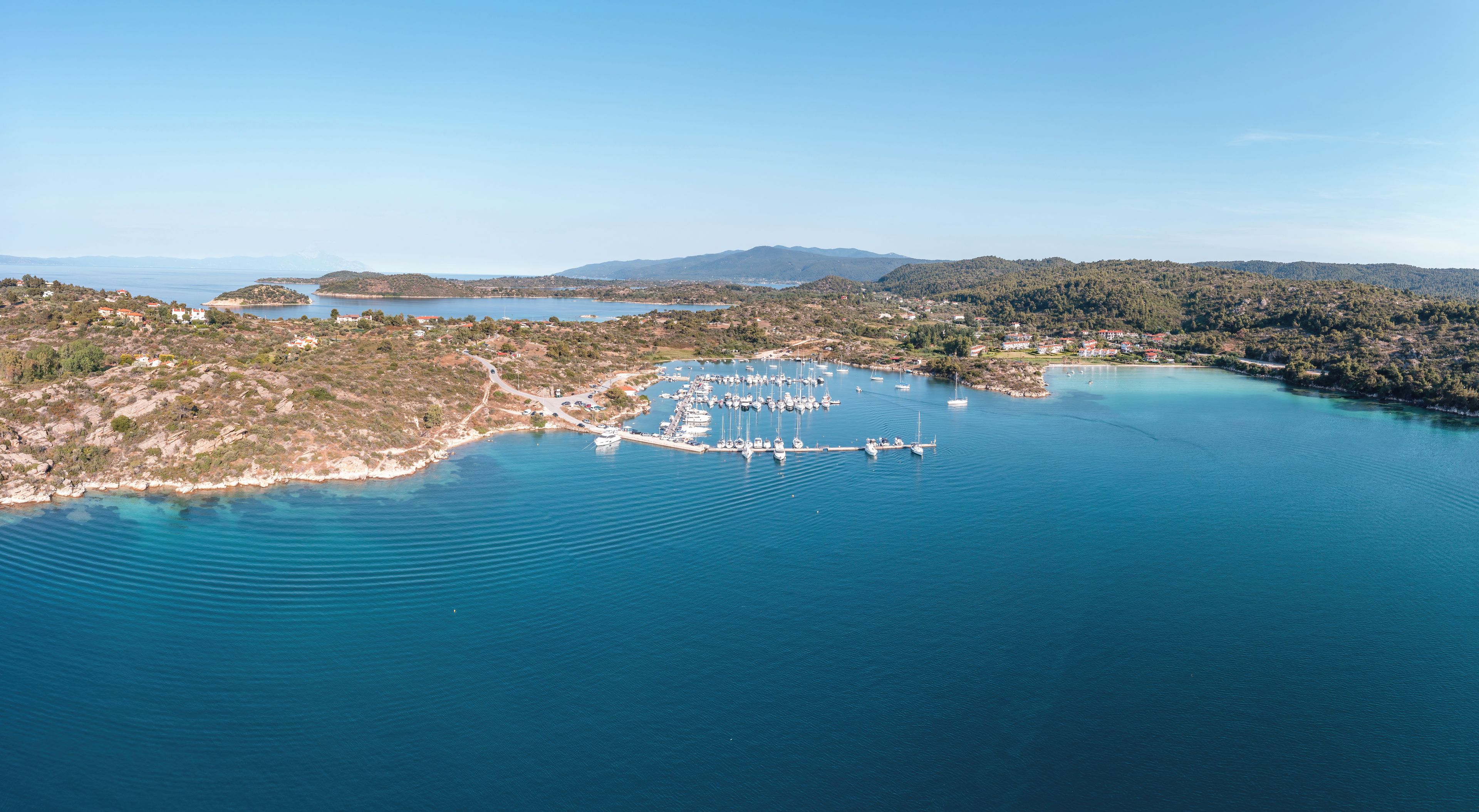 Vue aérienne d’une zone côtière avec un port de plaisance, des bateaux et des collines environnantes.