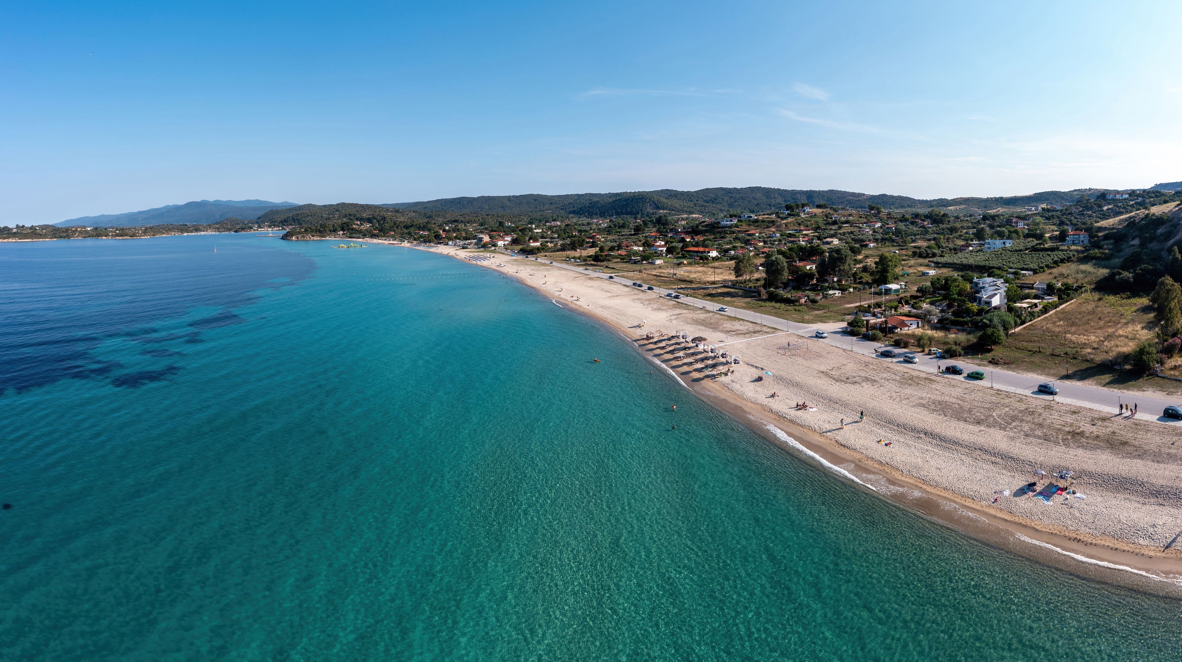 A coastal town with a beach, clear blue waters, and hilly terrain in the background.