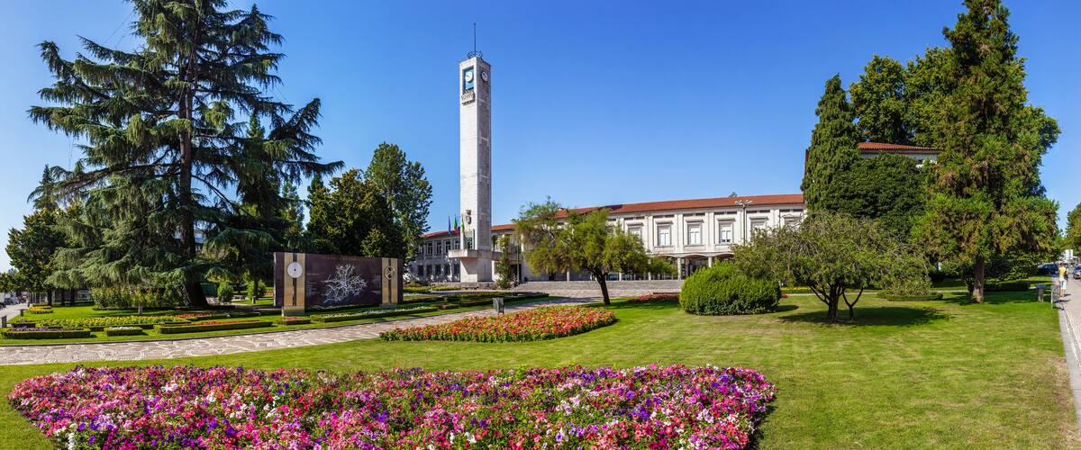 A park with a tall clock tower, a building with a red roof, and various trees and flowers.