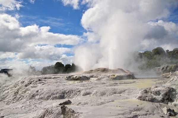 A geyser erupting in a geothermal area with rocky terrain and sparse vegetation.