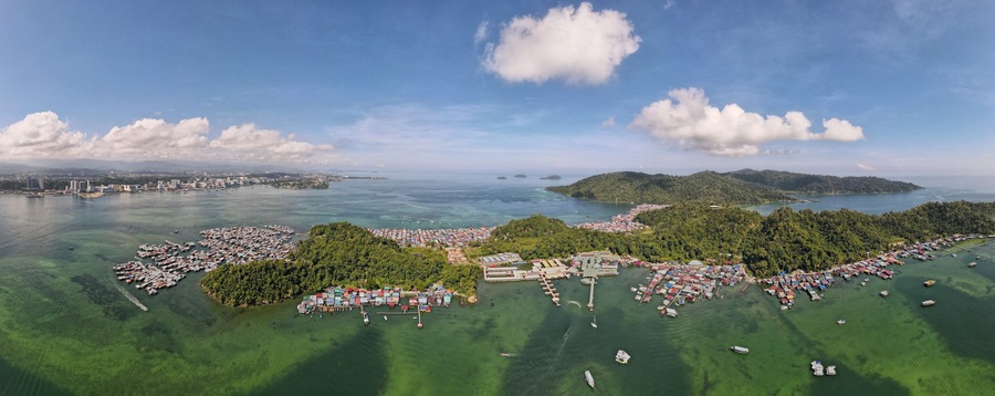 Vista aérea de un pueblo costero con un puerto deportivo, colinas verdes y cielo despejado.