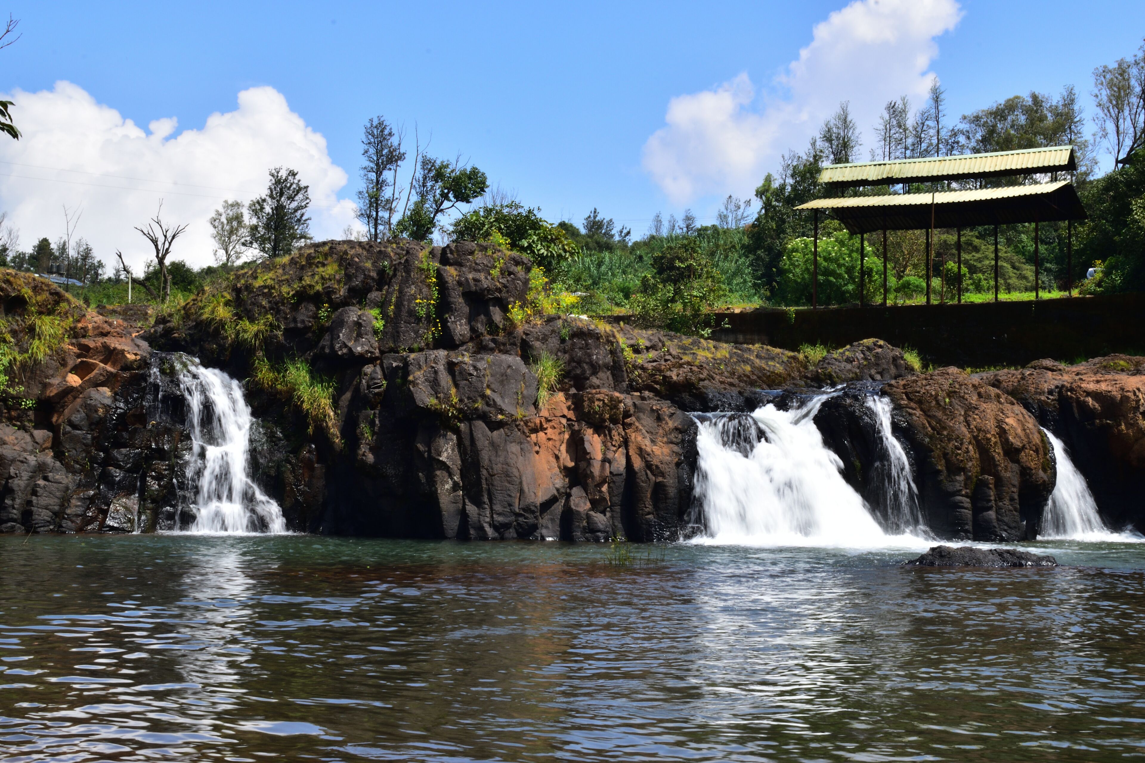 Uma cascata desliza sobre terreno rochoso em direção a um lago tranquilo, com uma construção coberta em uma colina ao fundo.