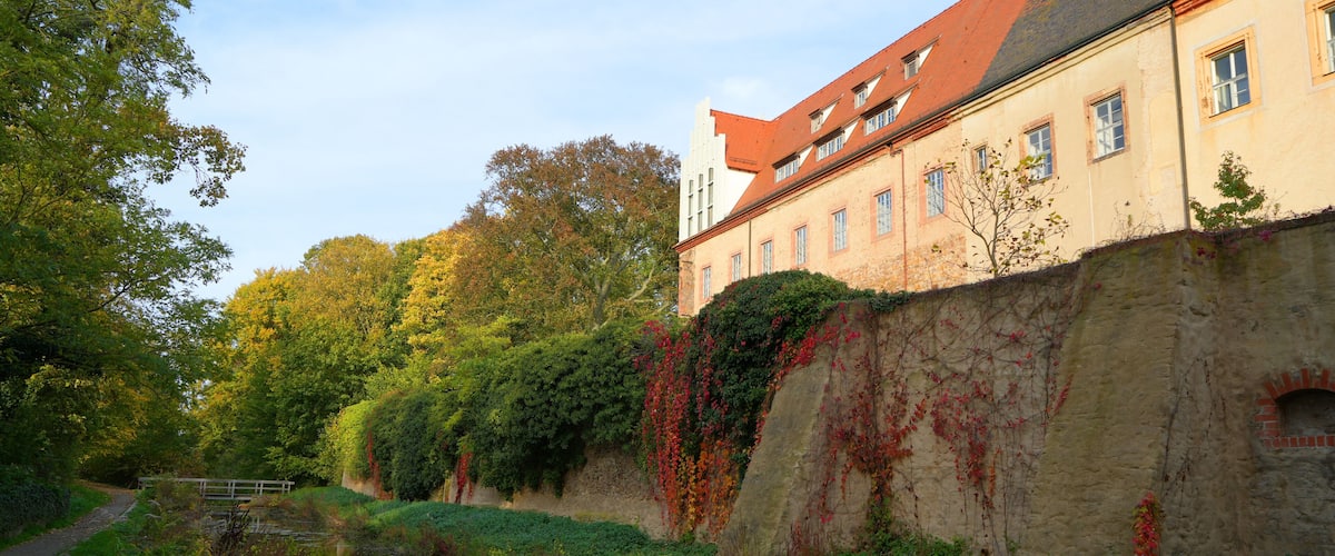 A historic building with a red roof, a river, and lush greenery.