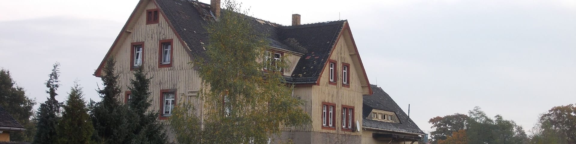 A large, two-story house with a sloped roof and a chimney, surrounded by trees and a railway track.