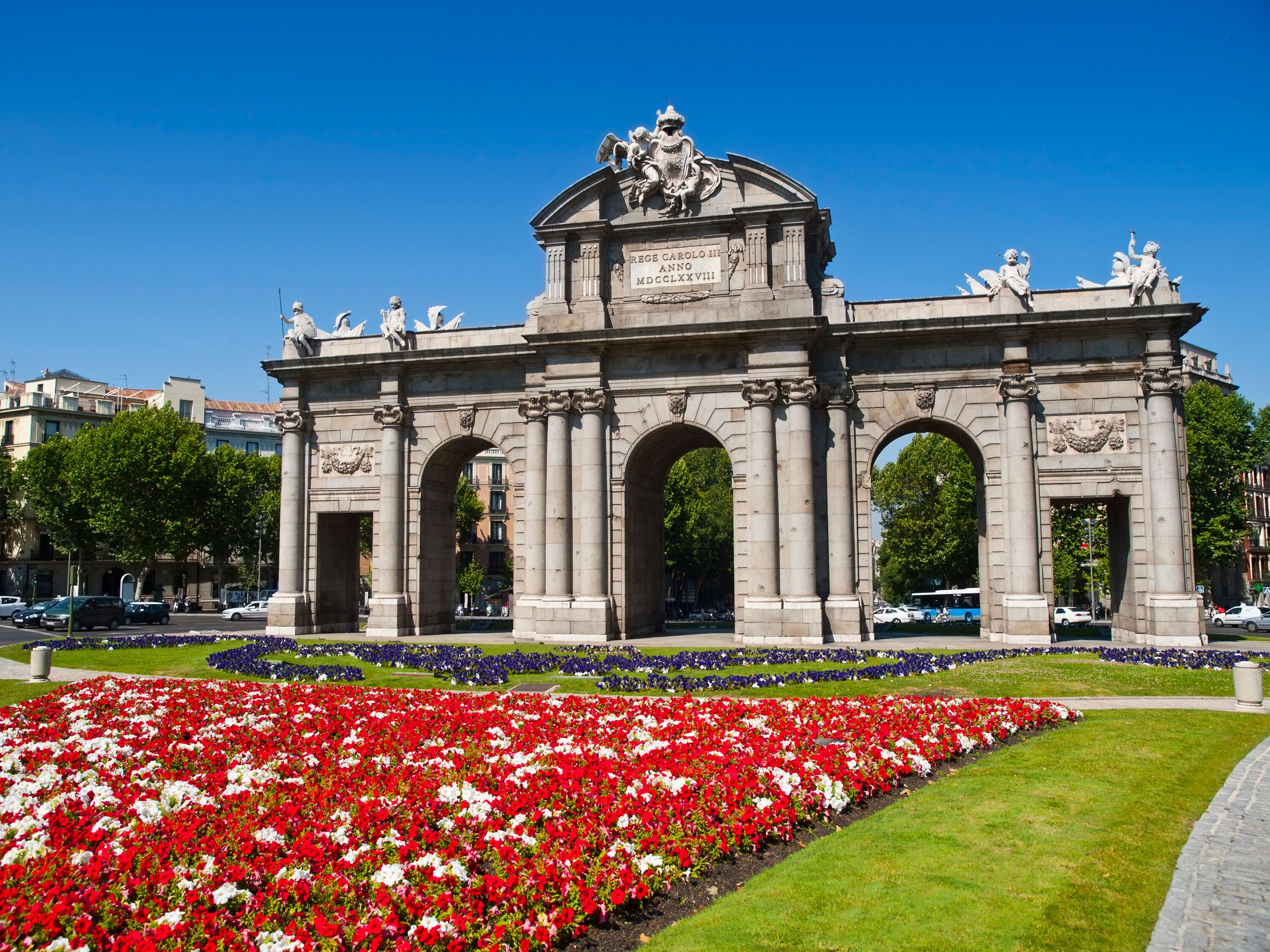 An ornate triumphal arch with statues and inscriptions, surrounded by a vibrant flower garden.