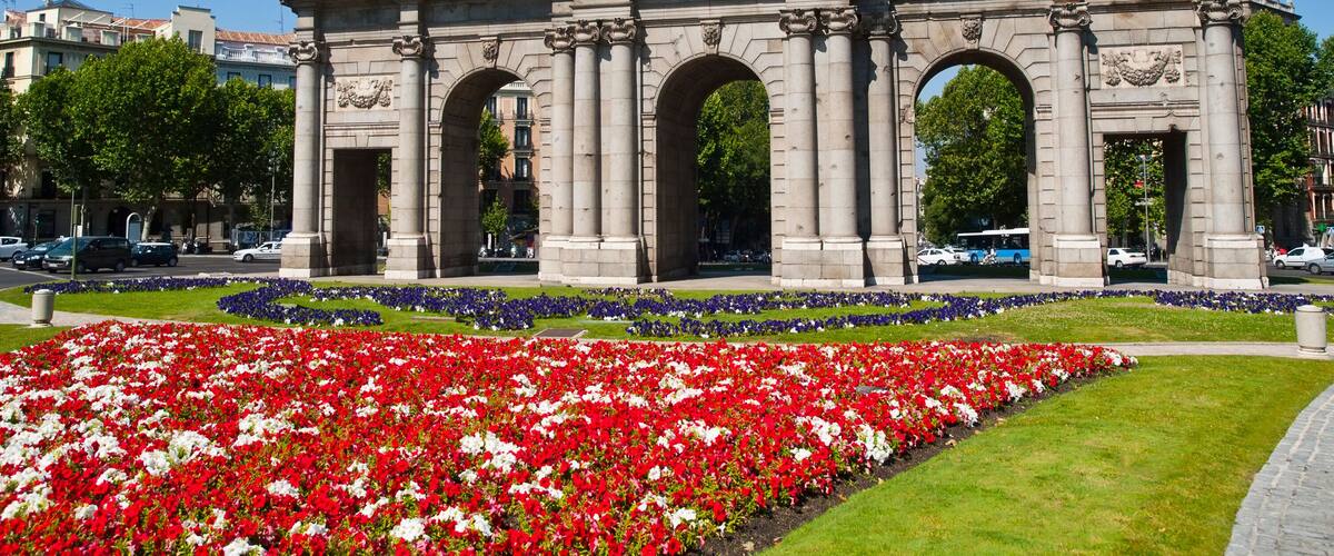 An ornate triumphal arch with statues and inscriptions, surrounded by a vibrant flower garden.