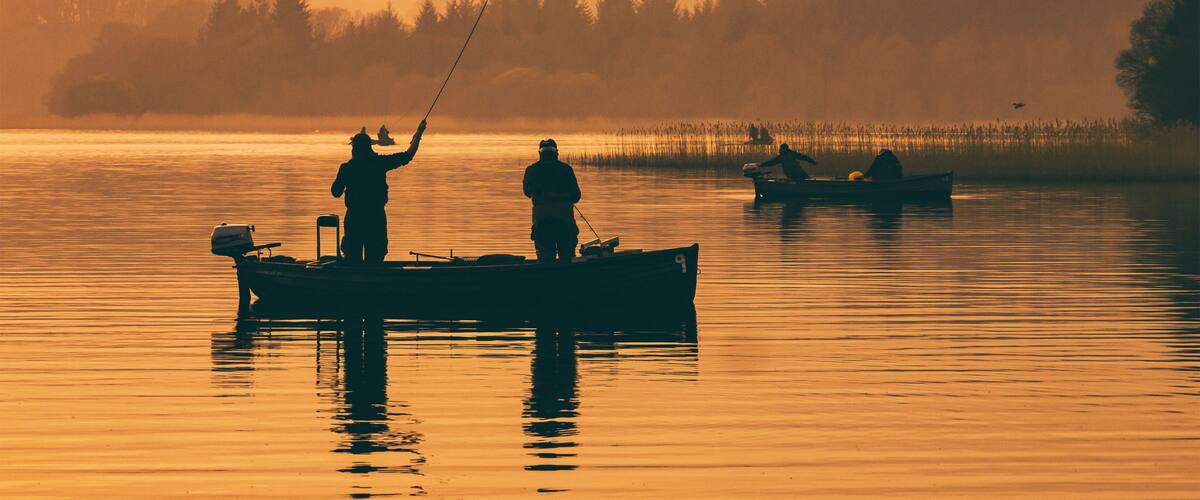 Um lago tranquilo ao amanhecer, com pescadores em barcos, rodeado por floresta densa e névoa.