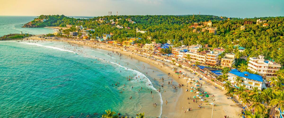 A coastal beach with clear turquoise waters, a sandy shore, and a backdrop of lush greenery and buildings.