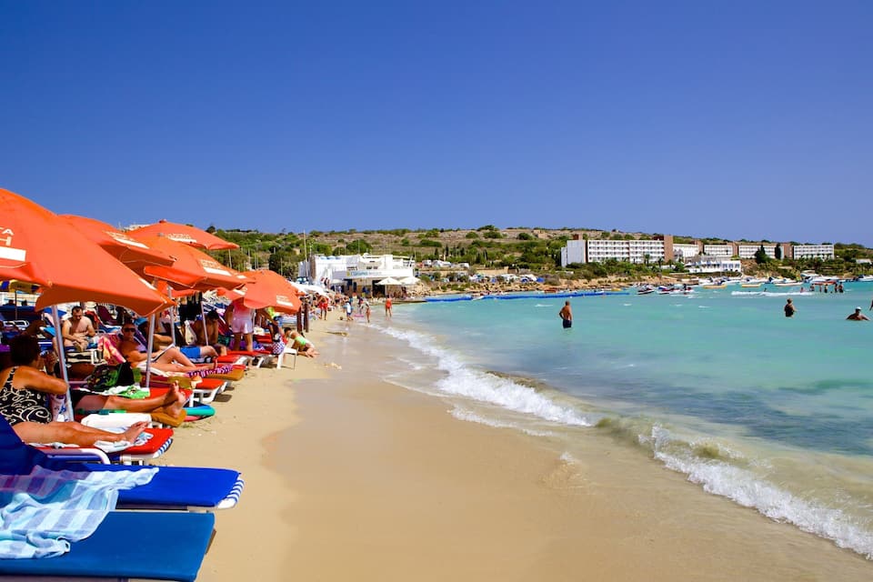 A beach with rows of sun loungers and umbrellas, clear turquoise water, and buildings in the background.