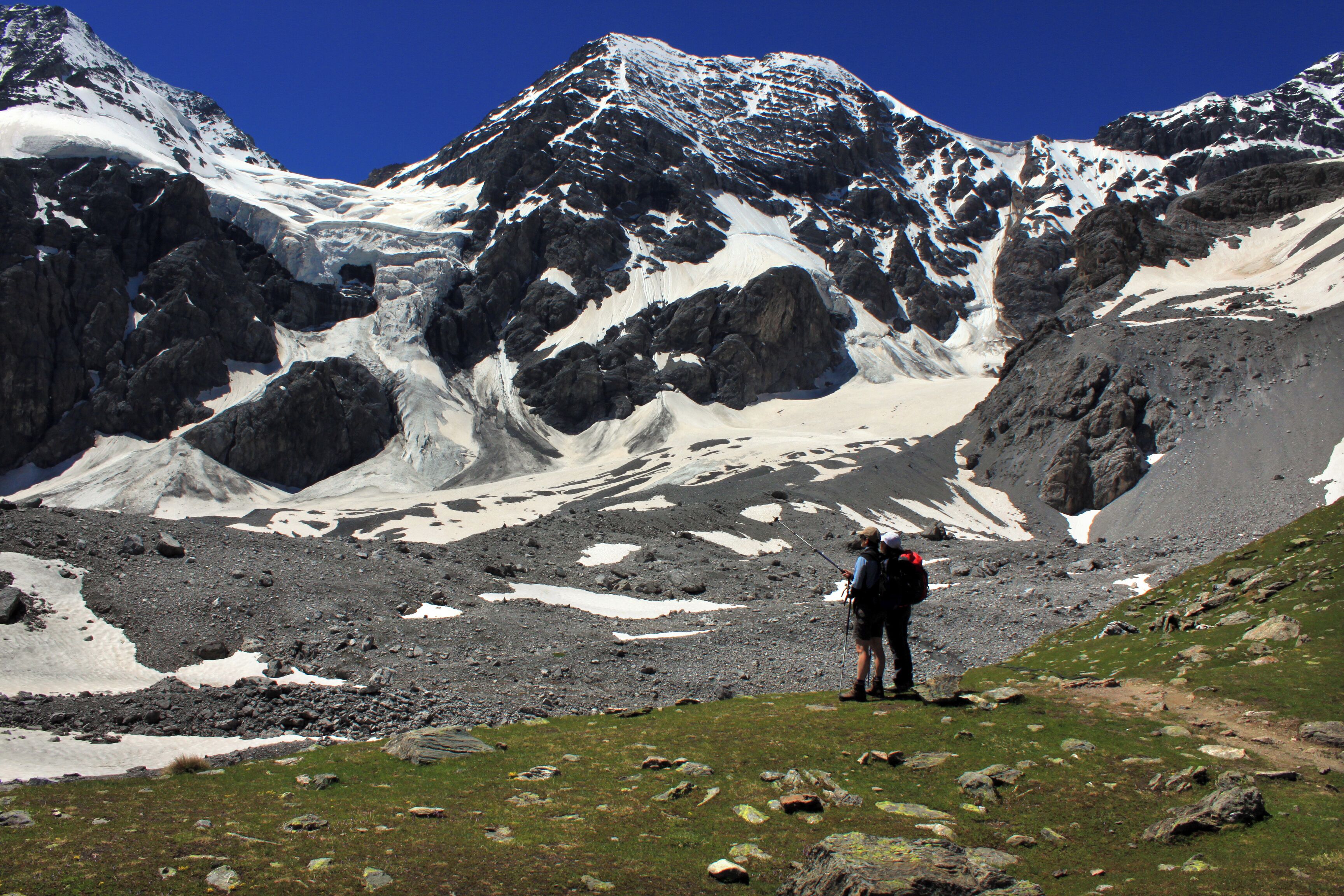 A mountainous landscape with snow-covered peaks and a hiker standing on a grassy slope.