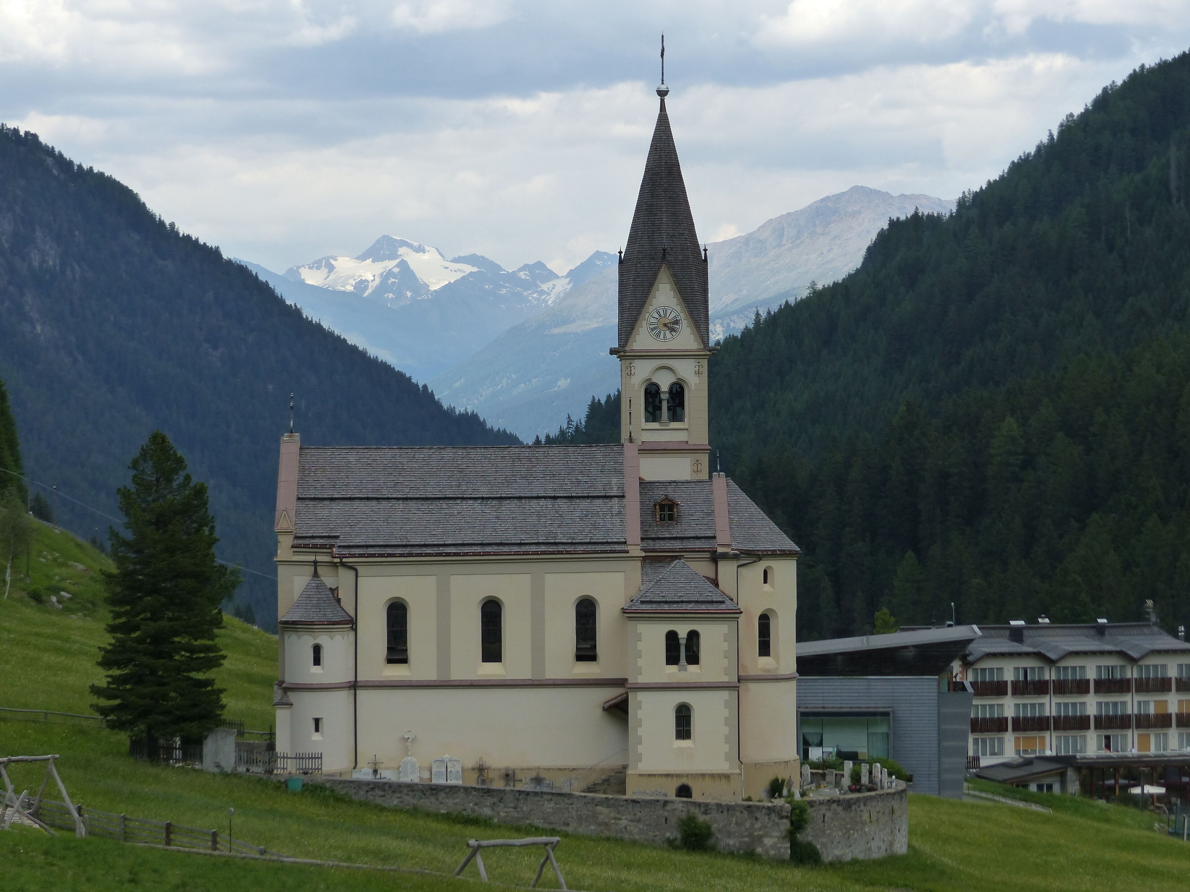 A church with a tall steeple and a clock tower, surrounded by mountains and a forested area.
