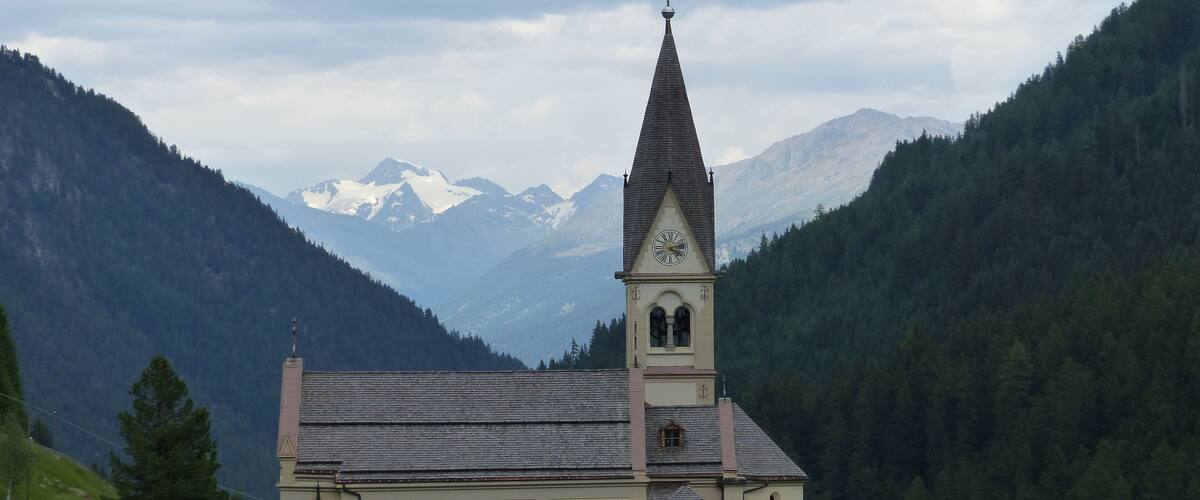 A church with a tall steeple and a clock tower, surrounded by mountains and a forested area.