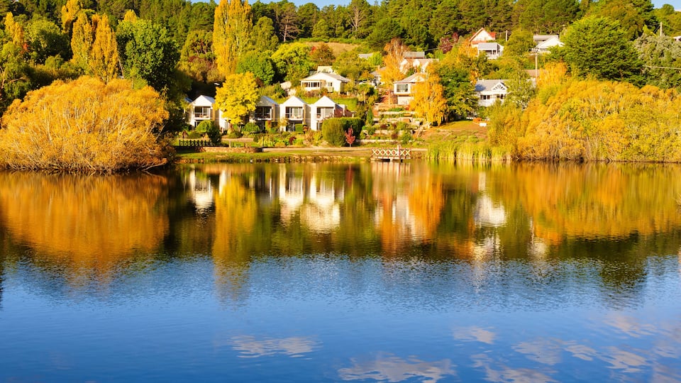 A serene lake with reflections of houses and trees, surrounded by autumn foliage.