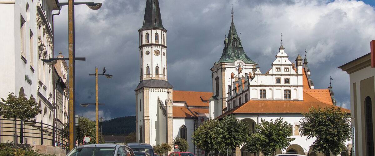 Una iglesia histórica con un alto campanario y una aguja más pequeña, rodeada de calles adoquinadas y coches estacionados.