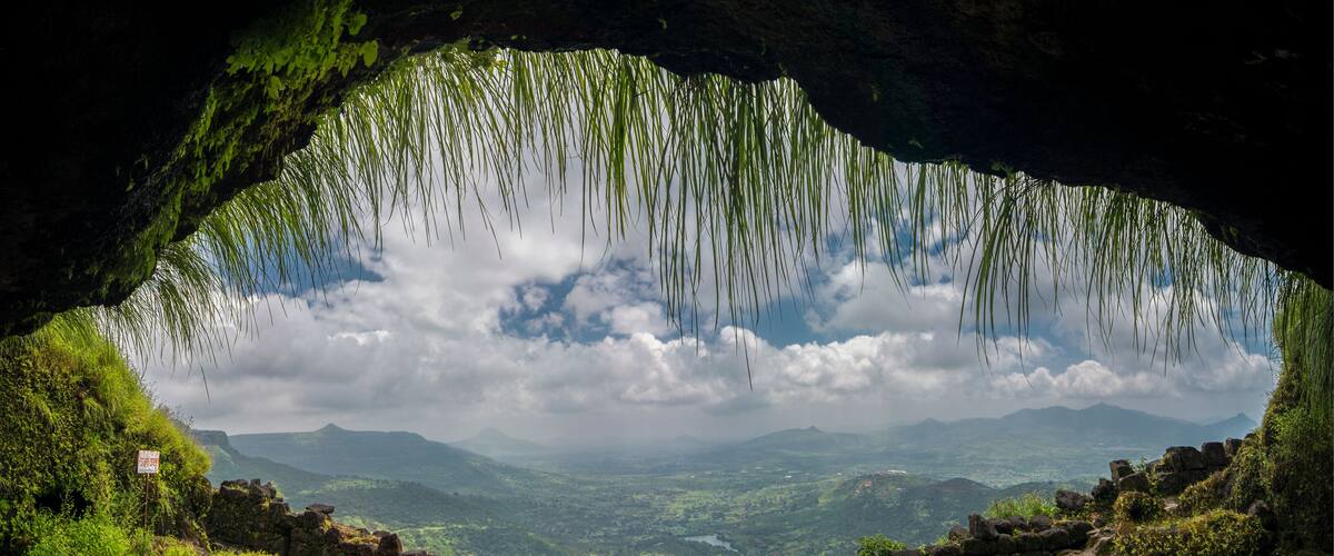 A cave entrance with hanging vegetation, a stone wall, and a view of distant mountains and valleys.