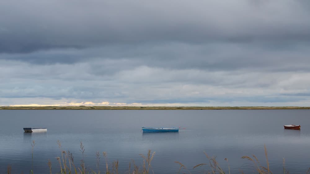 Ein ruhiger See mit drei Booten, bewölkter Himmel und fernen Dünen.