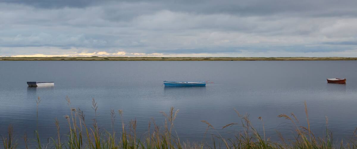 A calm lake with three boats, overcast sky, and distant dunes.