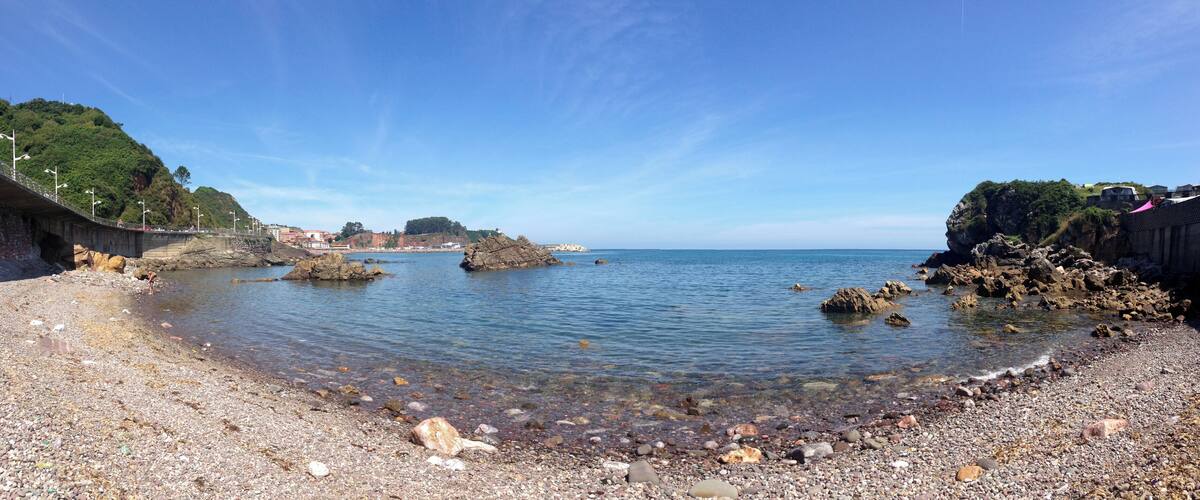 A pebble beach with clear water, rocky outcrops, and a distant town.