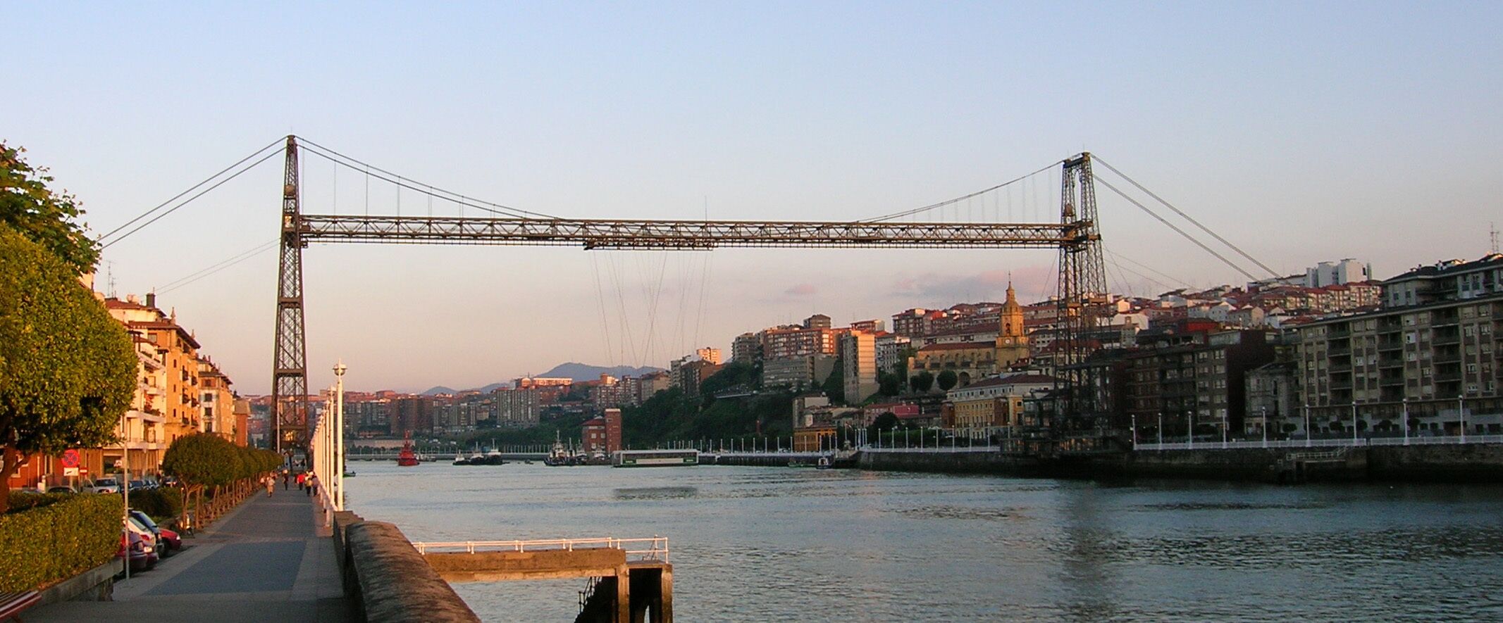 A large suspension bridge spanning a river with a cityscape in the background.