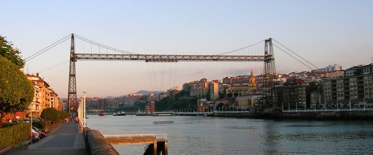 A large suspension bridge spanning a river with a cityscape in the background.