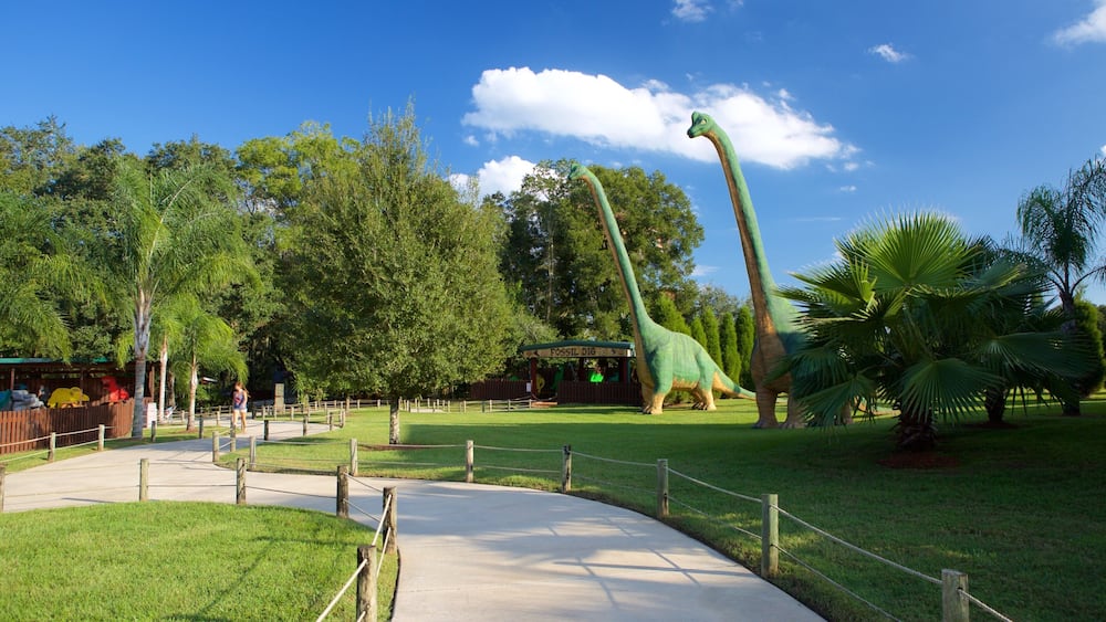 Une exposition sur les dinosaures comprenant une grande statue de dinosaure verte, un bâtiment consacré à la fouille de fossiles et un sentier délimité par une clôture.