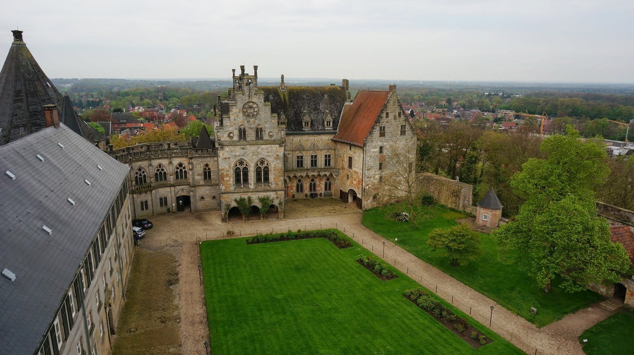 A historic castle with a large courtyard, surrounded by lush greenery and a view of the town in the background.
